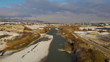 Flying-over-a-winter-landscape-and-towards-a-pedestrian-bridge-over-a-river-through-a-park---aerial-view