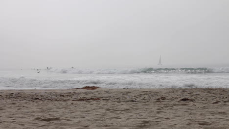 Marine-Layer-over-Venice-Beach,-CA-as-walkers-stroll-by-and-surfers-hit-the-waves