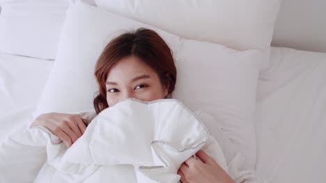 Beautiful-Asian-woman-smiling-under-a-blanket-on-a-white-mattress-on-the-morning-of-the-holiday