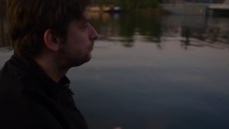 Slow-motion-side-profile-close-up-of-a-young-man-sitting-on-a-pier-of-a-reflective-lake-and-eating-potato-chips