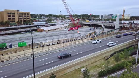 Slow-panning-low-aerial-drone-shot-of-busy-highway-exit-showing-bridge-overpass-intersection-with-vehicular-traffic,-road-construction-zone,-road-construction-equipment,-and-crane