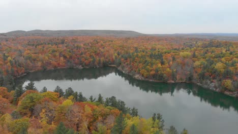 Sideways-aerial-footage-over-Pink-Lake-in-Gatineau-Quebec-with-the-mountains-in-the-background-and-orange-autumn-trees-all-over-the-place