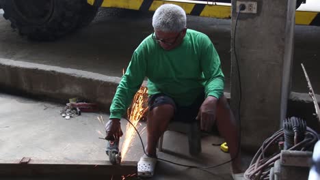 wideshot-of-an-old-man-with-white-hair-cutting-a-metal-bar-using-an-angle-grinder-with-no-protective-guards-like-face-shield,-gloves,-shoes-and-working-suit