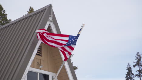 American-flag-mounted-on-an-old-a-frame-cabin-in-the-woods,-blowing-in-the-wind-slow-motion