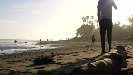 An-attractive-man-walking-on-the-beach-at-Rincon-point-in-California-at-sunset-with-people-and-palm-trees-in-silhouette-at-golden-hour