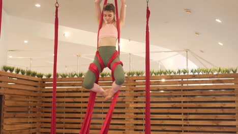 Young-brunette-woman-in-a-minimal-and-modern-gym-with-wooden-dividers-doing-flying-yoga-with-some-bright-red-hammocks,-doing-amazing-and-acrobatic-stunts