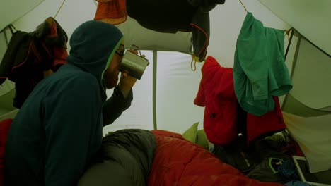 inside-the-tent-during-a-stormy-weather,-male-hiker-in-a-sleeping-bag-waiting-and-drinking-a-beverage,-tent-walls-are-shaking-in-a-gusts-of-wind,-wide-shot