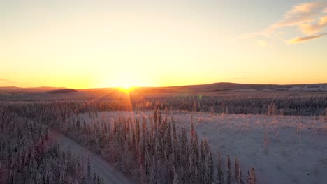 Aerial-view-of-sunset-over-snowy-frozen-road-in-deep-forest-with-hills-and-mountains