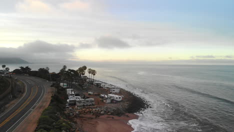 Aerial-drone-pulling-away-from-an-rv-campground-and-road-on-the-sand-beaches-of-Ventura,-California-with-ocean-waves-crashing-on-the-shore-at-sunrise