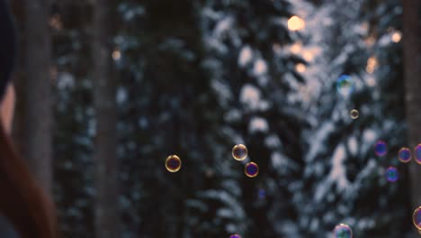 Brünette-Frau-Mit-Winterkleidung,-Die-Regenbogenfarbene-Seifenblasen-Bläst,-Die-In-Der-Luft-Schweben,-Mit-Einem-Verschneiten-Wald-Im-Hintergrund