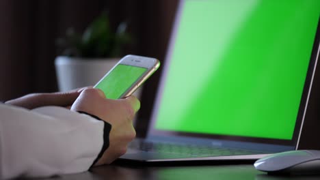 Holding-a-smartphone-with-green-screen,-scrolling-and-then-clicking-with-left-finger-on-the-upper-left-corner-of-the-screen-with-a-laptop-with-green-screen-in-the-back