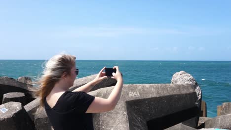 Young-woman-taking-photos-on-her-cellphone-of-the-ocean-on-a-pier-with-the-wind-blowing-her-hair-on-a-sunny-day