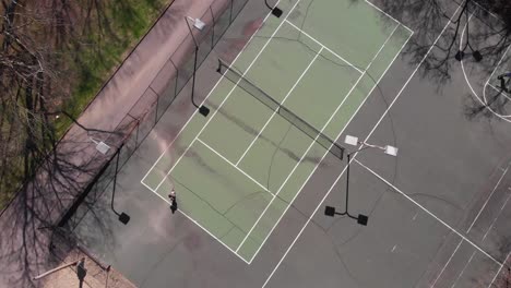 Drone-footage-looking-straight-down-while-flying-up-over-basketball-and-tennis-courts-with-someone-skateboarding-and-another-person-playing-basketball-with-shadows