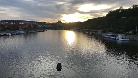 Jemand-Sitzt-In-Einem-Ruderboot,-Das-Allein-Mitten-Auf-Der-Moldau-Schwimmt,-Während-Im-Hintergrund-Die-Abendsonne-Hinter-Der-Prager-Burg-Untergeht