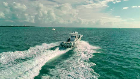 Luftaufnahme-Eines-Quad-Motor-Mittelkonsolenboots-Mit-Reisegeschwindigkeit-Und-Segelboot-Im-Hintergrund-In-Blauen-Küstengewässern