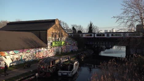 UK-February-2019--Three-trains-cross-each-other-on-bridge-over-Regents-canal-and-a-graffitied-wall-by-a-towpath-with-Canary-Wharf-financial-district-in-the-background