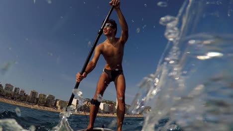 Young-man-standup-paddling-with-city-beach-in-the-background