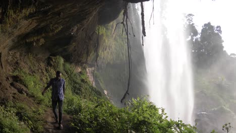 Slow-motion-shot-of-an-African-man-walking-in-a-cave-behind-a-tropical-waterfall