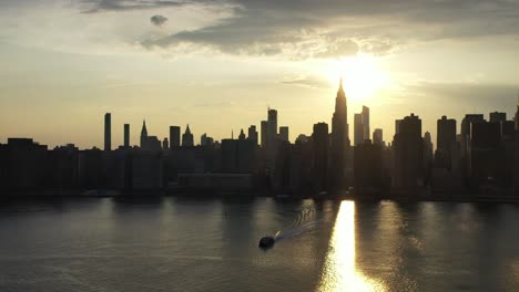 A-dolly-shot-moving-forward-towards-New-York-City---a-golden-sunset,-looking-down-at-the-river-as-a-ferry-moves-towards-the-drone-camera