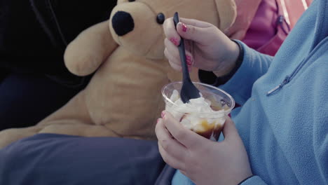 Close-up-of-little-girl-with-blue-jumper-nail-polish-and-teddy-bear-beside-her-holding-a-sundae-ice-cream-sitting-outdoor-4k