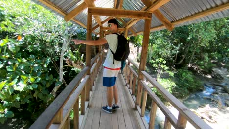 Un-Hombre-Que-Cruza-Un-Puente-De-Madera-Construido-A-Mano-Mientras-Señala-Algo-Fuera-De-Cámara-Mientras-Se-Encuentra-En-La-Exuberante-Jungla-Tropical-De-Bohol,-Filipinas