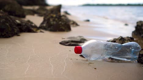 Plastic-bottle-sits-motionless-amongst-rocks-on-small-sandy-beach,-water-in-background