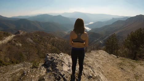 Atlete-girl-standing-at-a-top-of-a-mountain-making-hair-ponytail-with-beautiful-canyon-lake-in-front-of-her