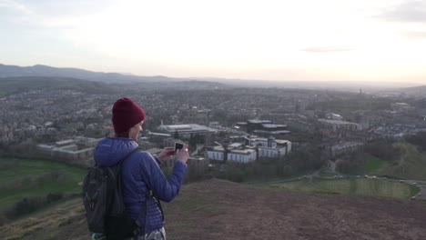Circling-parallax-slow-motion-shot-of-girl-taking-a-photo-with-smartphone-from-the-Arthurs-seat-mountain-in-evening-with-city-of-Edinburgh-in-the-background-during-wonderful-golden-hour-with-sunset
