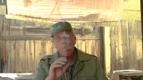 Portrait-of-a-mature-male-in-his-60s-smoking-a-cigar,-sitting-outdoors-under-a-shade-cloth
