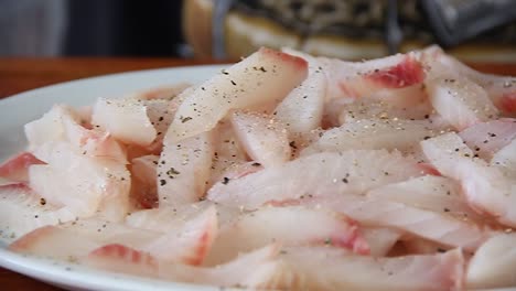 Raw-white-fish-on-a-white-plate-being-seasoned-with-salt-and-pepper-in-slow-motion-with-slider-moving-sideways