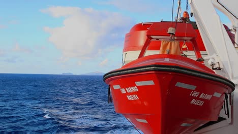 Red-safety-emergency-lifeboat-attached-to-ferry-passenger-transportation-ship-navigating-through-the-ocean,-blue-sky-with-clouds