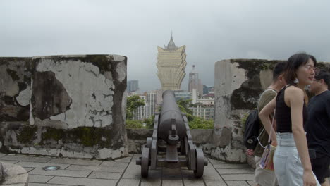 Tourists-passing-by-the-old-metal-cannon-on-top-of-Fortaleza-do-Monte-,-pointing-in-the-direction-of-Hotel-Grand-Lisboa-in-Macau,-Macau-SAR,-China