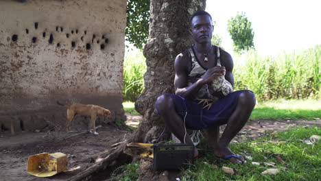 Young-man-sitting-on-a-small-chair-while-petting-a-chicken-and-listening-to-the-radio-with-dog-passing-by-in-the-background,-Uganda-Africa