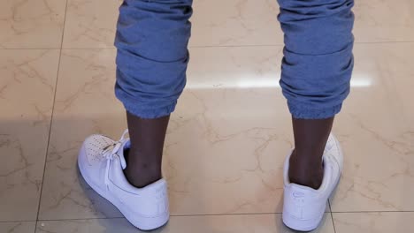 Close-up-of-a-black-Male-model-legs-with-rolled-up-hem-of-his-blue-pants-wearing-white-sneakers-doing-a-360-degree-turn-on-a-beige-floor---interior-scene-tripod-shot