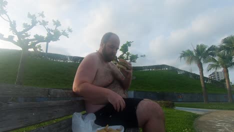 Obese-man-eating-a-greasy-burger-while-exposing-his-fat,-ugly-stomach-sitting-on-public-park-bench