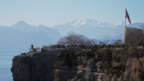 Totale-Aufnahme,-Tourist-Auf-Den-Klippen-Von-Antalya,-Der-Herumläuft,-Malerische-Aussicht-Auf-Die-Toros-Berge-Im-Hintergrund,-Türkei-Flagge-An-Der-Seite