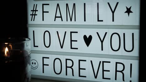 A-lit-glass-candle-in-front-of-a-billboard-with-the-text-Family-Love-You-Forever,-a-hashtag-and-some-symbols-
