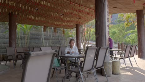 Chinese-elderly-male-sitting-in-outdoor-seating-area-under-roofing-reading-book-alone