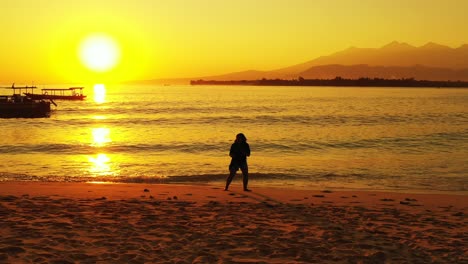 Romantic-girl-walking-on-empty-quiet-exotic-beach-at-sunset-with-glowing-yellow-sky-reflecting-on-peaceful-sea-surface,-Indonesia