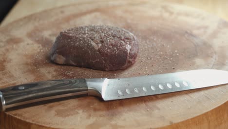 Slow-Motion-Slider-Shot-of-Seasoning-Steak-With-Salt-Then-Patting-it-Down-in-Kitchen-Next-to-a-Sharp-Chefs-Knife-on-a-Thick-Wooden-Chopping-board