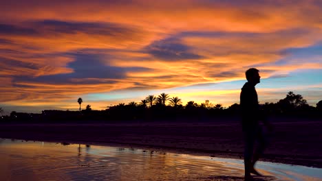 Eine-Person,-Die-Von-Rechts-Nach-Links-über-Das-Wasser-Geht-Und-Den-Atemberaubenden-Sonnenuntergang-An-Einem-Wunderschönen-Tropischen-Strand,-Wolken-Und-Erstaunlichen-Farben-Beobachtet,-Ist-Erstaunt