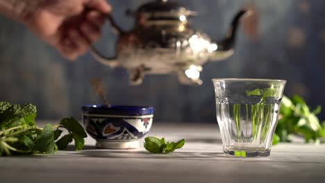 Weitwinkelaufnahme-In-Zeitlupe,-In-Der-Eine-Hand-Eine-Verzierte-Teekanne-In-Die-Hand-Nimmt-Und-Tee-In-Ein-Glas-Mit-Marokkanischer-Schüssel-Und-Minze-Gießt