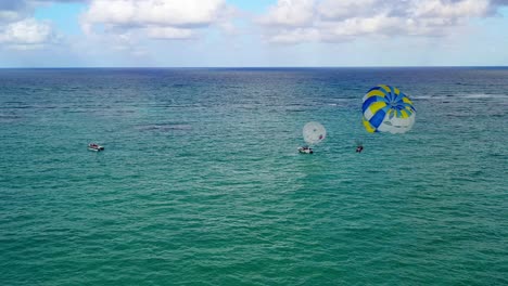 Two-people-parasailing-together-in-yellow-and-blue-parachute-in-open-green-ocean-waters,-circle-aerial
