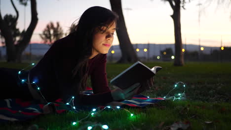 A-cute-young-woman-reading-a-fictional-story-book-outdoors-at-twilight-with-lights-glowing-SLIDE-RIGHT