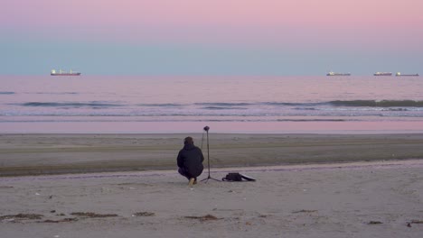 The-crouching-sound-technician-is-recording-environments-on-the-beach-at-sunset,-the-pink-sky-and-the-waves-of-the-sea-with-the-horizon-in-the-background-of-the-sequence