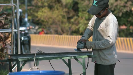 Medium-Slow-Motion-Shot-of-an-Asian-Man-at-the-Side-of-the-Road-Using-an-Angle-Grinder-on-a-Piece-of-Metal