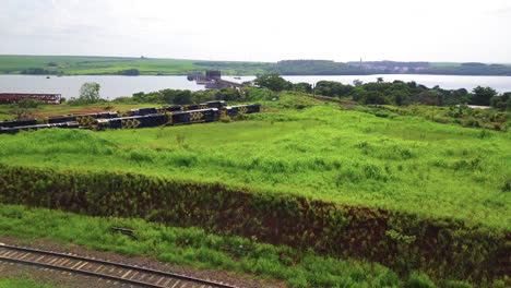 Stationary-train-wagons-waiting-to-load,-beautiful-green-vegetation-all-around,-in-the-background-a-beautiful-river,-a-railroad-bridge-and-boats-docked-on-the-coast