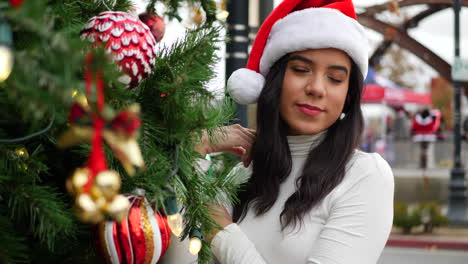 Eine-Fröhliche-Frau-Mit-Festlichem-Weihnachtsmannhut-Schmückt-Einen-Weihnachtsbaum-Mit-Ornamenten,-Glöckchen-Und-Lichtern