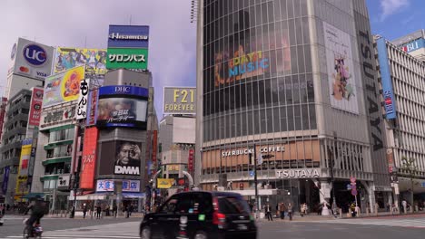 Vehículos-De-Transporte-Conduciendo-En-El-Cruce-De-Shibuya-En-Tokio,-Japón,-Con-Anuncios-Digitales-En-La-Pared-Exterior-De-Los-Edificios-Comerciales-Durante-El-Día---Plano-General