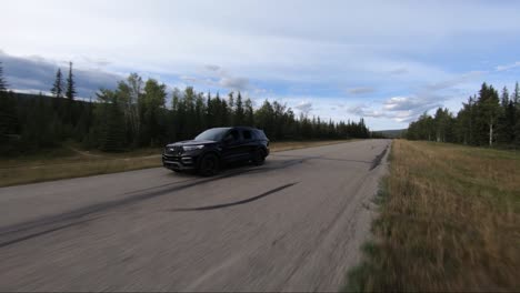 A-black-suv-driving-towards-a-drone-flying-in-front-of-it-on-a-country-road